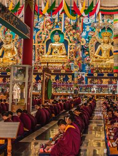 many buddhist monks are sitting at their desks in a large room with colorful banners hanging from the ceiling