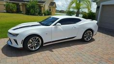 a white camaro is parked in front of a house with palm trees and grass