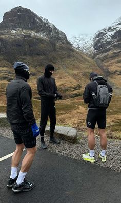 three men standing on the side of a road with mountains in the backgroud