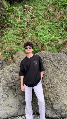 a young man standing in front of a rock and grass covered hill with trees on it