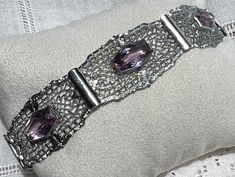 "This hard-to-find Ostby & Barton bracelet dates from the 1930s.  In 1879 Engelhart Ostby formed a partnership with Nathan Barton. The firm became the world's largest producer of gold rings. In 1912 he decided to return from one of his many European trips on the Titanic, carrying with him his old doctor's bag he always used when travelling, filled with gems, precious stones, and other valuables that he had bought in Paris. Unfortunately, he never made it back, as he went down with the Titanic. The business continued into the 1950s. The sterling and paste bracelet is composed of six filigree panels connected by bars with connector links hidden on the back.  Each panel is centered with a long hexagonal purple crystal paste.  In open-back settings, the sparkly stones are bezel-set with fine s Silver Art Deco Bracelets With Hand Set Details, Adjustable Silver Art Deco Bracelets, Formal Art Nouveau Amethyst Jewelry, European Trips, Victorian Hallmarked Silver Bracelets, Luxury Victorian Hallmarked Sterling Silver Bracelet, Filigree Bracelet, The Titanic, Doctor Bag