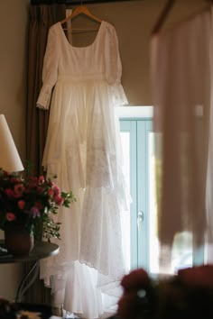 a wedding dress hanging on a hanger in front of a window with curtains and flowers