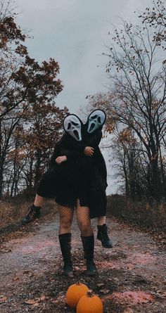 two people in costumes standing on a dirt road with pumpkins and trees behind them