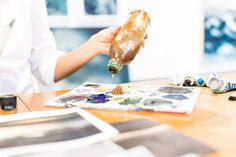 a person is holding a bottle with some paint and other items on the table next to it