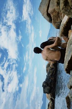 a man sitting on top of a rock next to the ocean under a cloudy blue sky