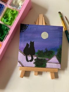 an easel with some paint on it next to a painting and a cat sitting on the roof