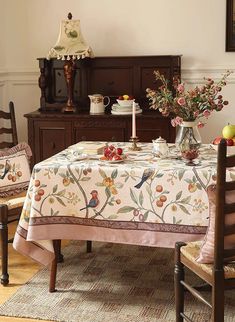 a dining room table covered with a white and floral tablecloth next to two chairs