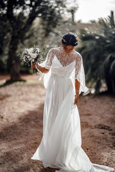 a woman in a white dress is walking down the dirt road with her wedding bouquet