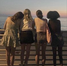 four girls are standing on a fence looking at the water and sunset over the ocean