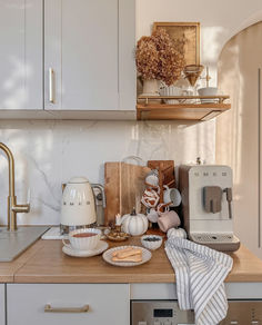 Cozy kitchen setup featuring cream-colored Smeg appliances, including an electric kettle and espresso machine, on a wooden countertop. The decor includes dried hydrangeas in a vase, framed artwork, wooden cutting boards, and ceramic mugs. A cup of coffee with milk, a plate of ladyfingers, and a bowl of blueberries add a warm, inviting touch. A striped kitchen towel drapes over the counter, creating a charming and relaxed atmosphere. Smeg Kitchen Appliances, Modern Cottage Kitchen, Naturalist Decor, Estilo Cottage, Girl Apartment Decor, Coffee Nook, Modern Cottage