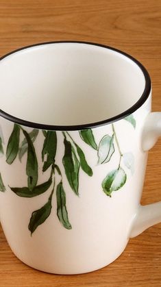 a white coffee cup with green leaves painted on the side and black rim, sitting on a wooden table