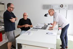 three men are standing around a table with papers on it and one man is pointing at something