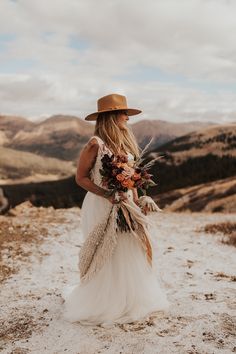 a woman in a white dress and brown hat is holding a bouquet on the top of a mountain