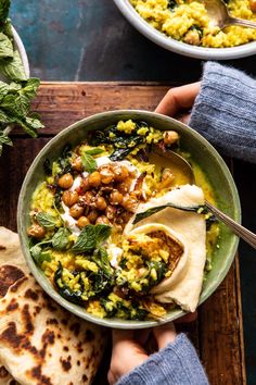 two hands holding a bowl of food with pita bread and greens on the side