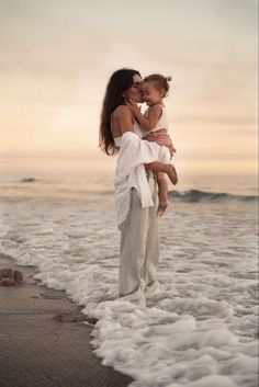 a woman holding a small child on the beach at sunset with foamy water around her