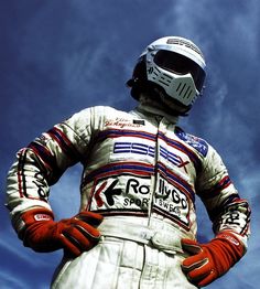 a man in a racing suit and helmet standing with his hands on his hips against a blue sky