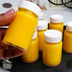 a hand holding a jar filled with yellow liquid next to other jars on a table