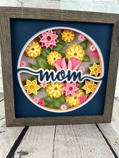 the word mom is surrounded by flowers in a wooden frame on a wood table with a blue background