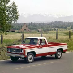 a red and white pickup truck driving down the road in front of a farm house