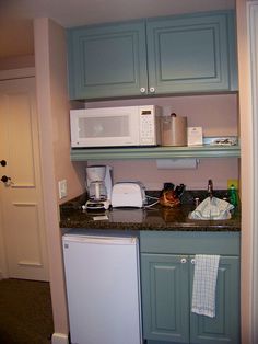 a small kitchen with blue cabinets and white appliances on top of the countertop, in front of a doorway that leads to another room