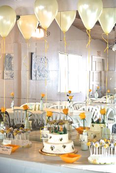 a table topped with lots of cake covered in white frosting and yellow balloons hanging from the ceiling