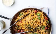 a skillet filled with pasta and vegetables on top of a white table next to two spoons