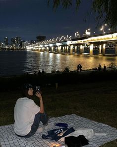 a man sitting on top of a blanket next to a river under a bridge at night