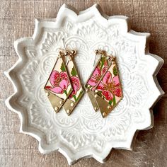 pink and gold earrings with floral designs on them sitting on a white plate in front of a wooden table