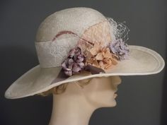 "Wide brim straw with a bouquet of flowers and veiling. Perfect for a garden party or Kentucky Derby. Off white straw has a 2.5\" white embossed ribbon band at crown. Flowers in shades of beige/mauve/purple, lilac. Mauve ribbon detail with white veiling. Mauve biot feathers. Trimmed in white grosgrain ribbon. Millinery wired brim to hold shape. Fit 22.5\" to 23\"" Summer Wedding Cloche Hat, Beige Straw Hat For Garden Party, Vintage Beige Fascinator For Evening, Cream Fascinator Hat For Garden Party, Beige Vintage Fascinator For Royal Ascot, Summer Wedding Brimmed Cloche Hat, Summer Wedding Cloche Hat With Brim, Spring Wedding Cloche Hat, Beige Cloche Hat For Wedding