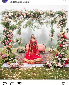 a woman in a red and gold wedding outfit standing under an arch with flowers on it