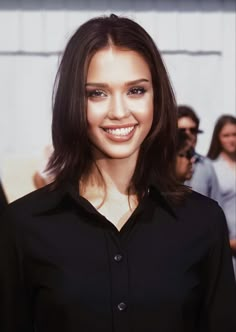 a woman with long brown hair and black shirt smiling at the camera while standing in front of a group of people