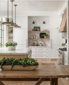 a kitchen with white cabinets and wooden table in the foreground is an oven, countertop, sink, and potted plants