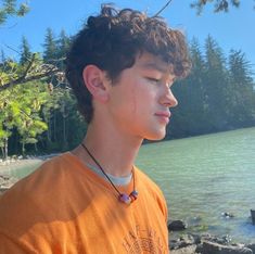 a young man wearing an orange shirt standing next to the water with trees in the background