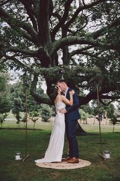 a bride and groom kissing under a large tree at their outdoor wedding ceremony in the park