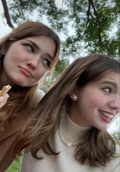 two young women are posing for the camera and eating food together in front of some trees