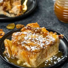 a piece of cake on a black plate covered in powdered sugar next to a jar of honey