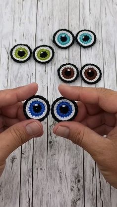 two hands are holding small crocheted eyeballs in front of them on a wooden table