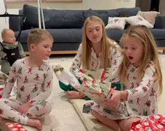 three children sitting on the floor in matching christmas pajamas and looking at an open present