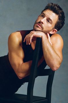 a man sitting on top of a black chair next to a gray wall with his arms crossed