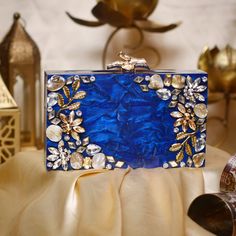 Overview Make a stunning statement on your special day with this luxurious blue acrylic bridal clutch. Crafted with exquisite detail, this minaudiere features a rich blue hue complemented by intricate gold floral accents and dazzling crystal embellishments. The structured design and elegant gold clasp closure make it a chic and practical accessory for brides, bridesmaids, or any evening event. Each clutch is carefully handcrafted, showcasing a combination of modern resin artistry with timeless g Luxury Blue Rectangular Clutch, Glamorous Blue Rectangular Clutch, Elegant Blue Clutch Ideal As A Gift, Blue Glamorous Party Clutch, Formal Handmade Blue Clutch, Blue Rectangular Evening Bag For Wedding, Elegant Blue Clutch For Wedding Guest, Blue Elegant Clutch For Wedding Guest, Blue Luxury Evening Bag As Gift