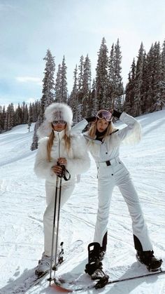 two women in white snowsuits posing for a photo on skis with trees in the background