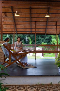 two women sitting at a table under a covered area with plants and trees in the background