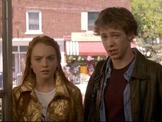 two young people standing next to each other in front of a window with buildings behind them