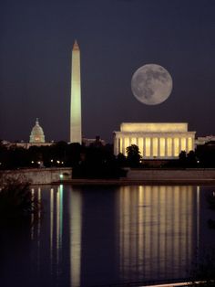 the washington monument is lit up at night with the moon in the sky above it