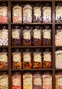 many jars filled with different types of candies on display in a wooden shelf next to each other