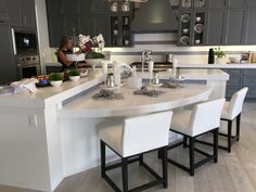 a woman is sitting at the counter in a large kitchen with gray cabinets and white chairs