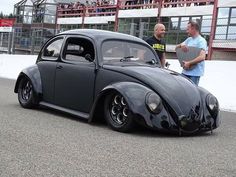 two men standing next to a black vw bug