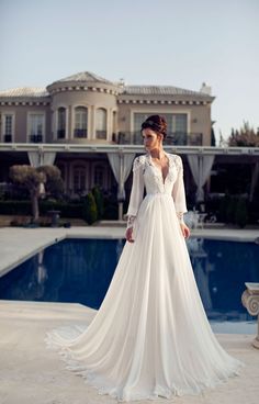 a woman standing in front of a swimming pool wearing a white wedding dress with long sleeves