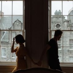 two people standing in front of a window looking out at the city below them and holding hands with each other