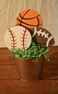 an arrangement of sports balls in a pail with greenery on the table next to it